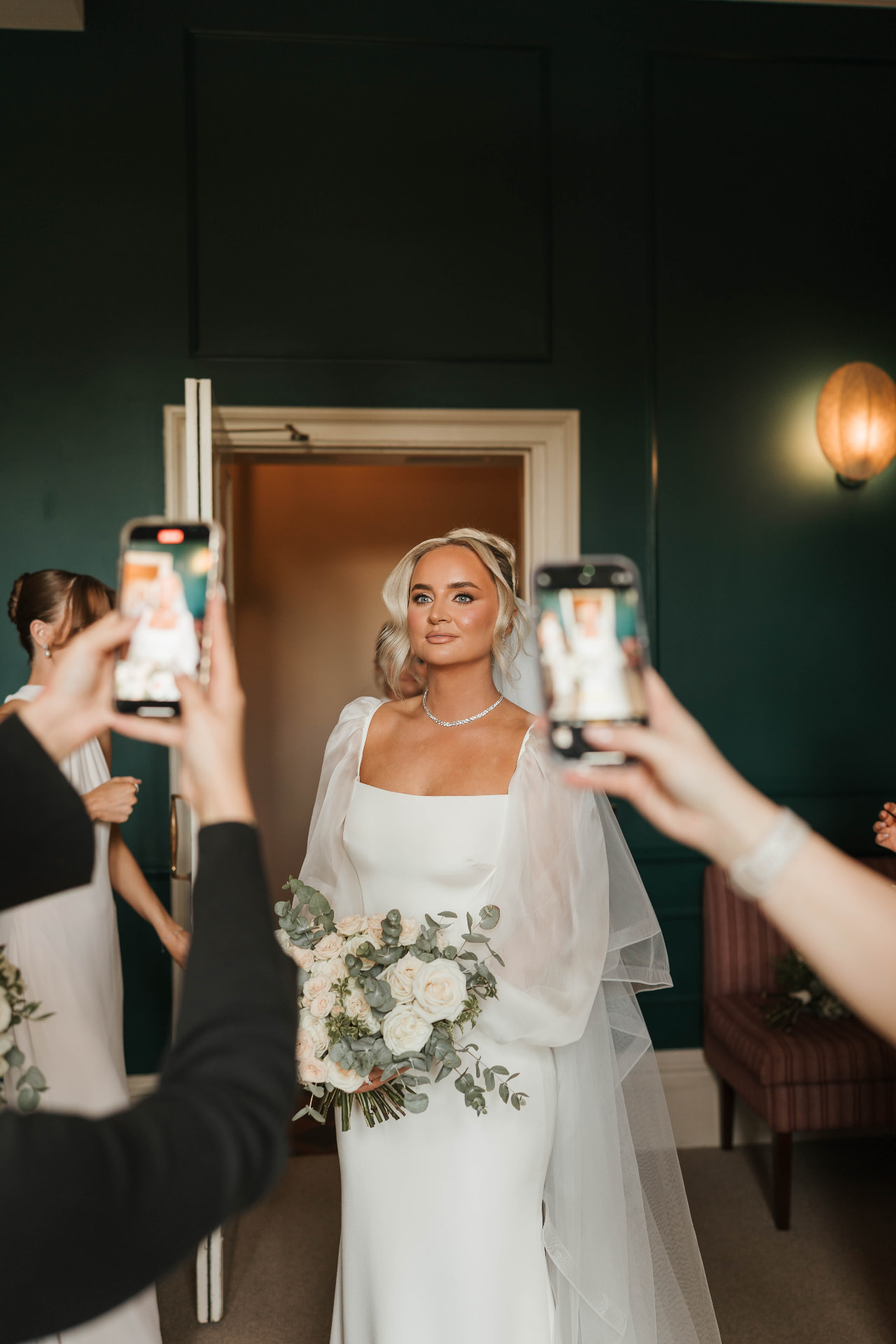 Bride holding a bouquet of ivory and blush roses, framed by guests holding up phones