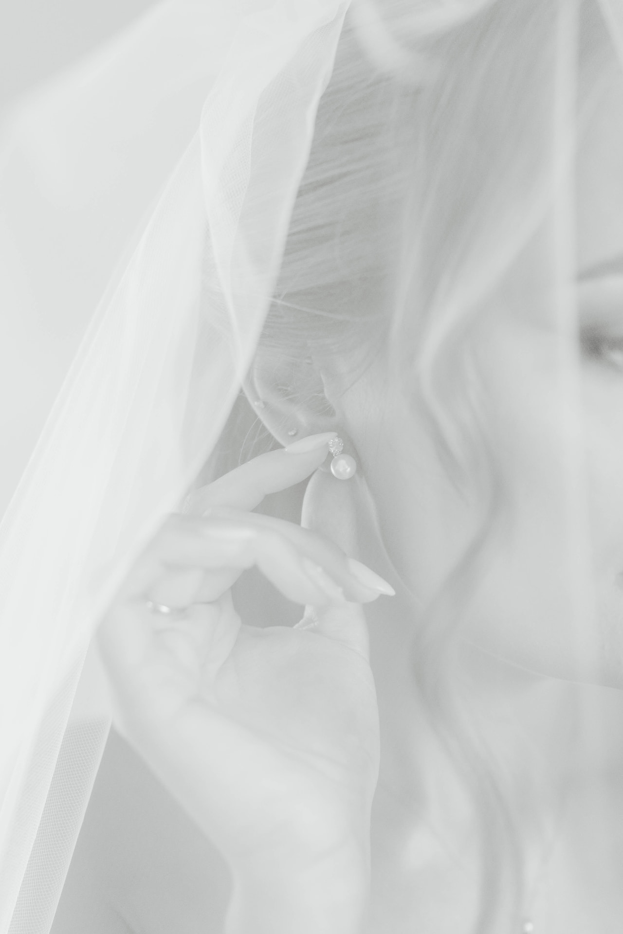 Bride in a sheer tulle veil touching a pearl drop earring, in black and white